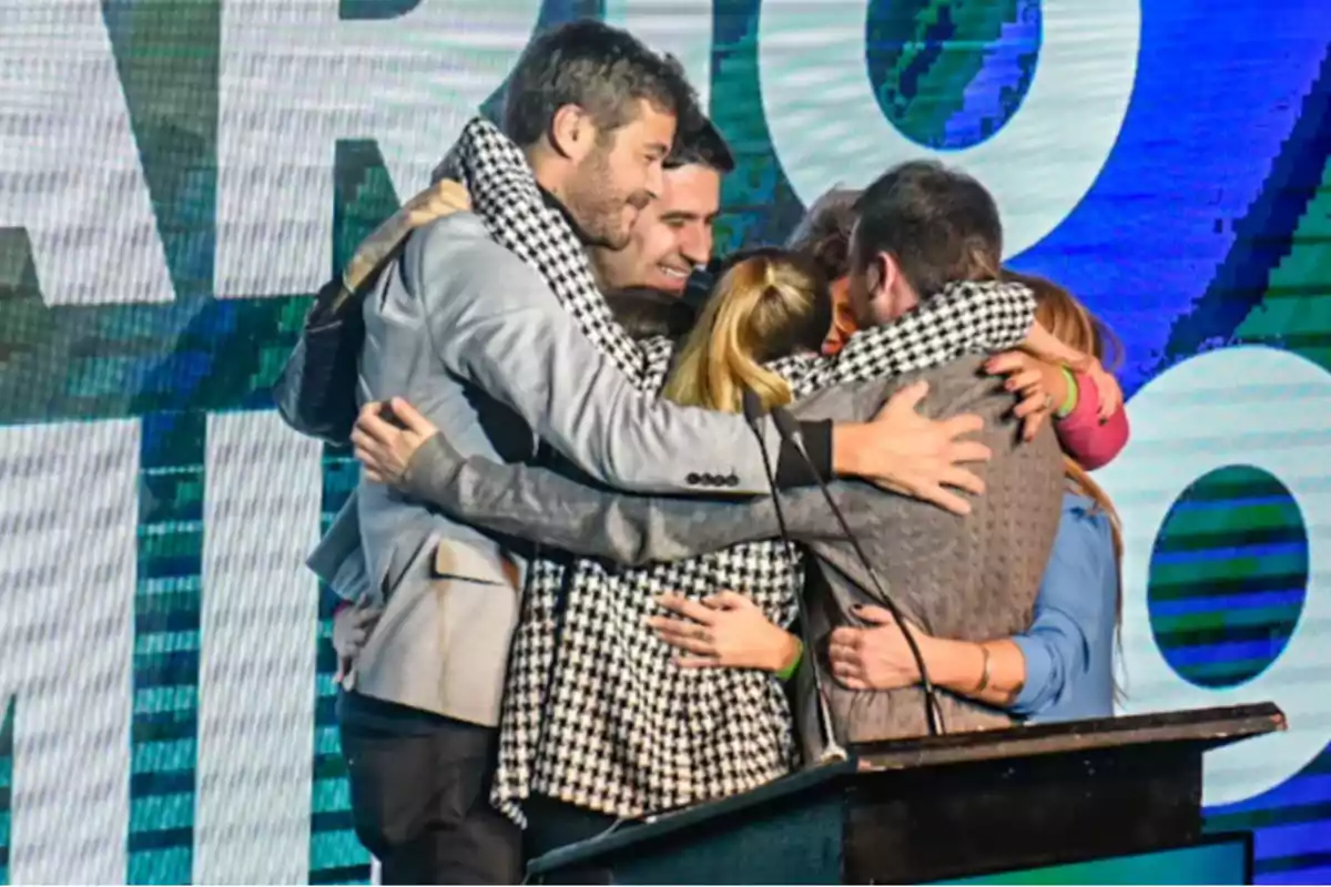 Grupo de personas abrazándose con alegría sobre un escenario frente a una pantalla grande