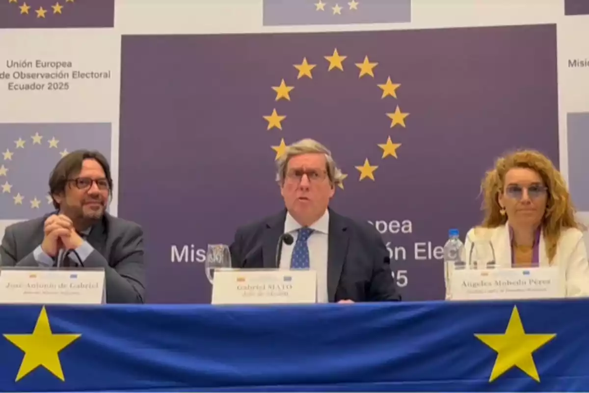 Three people seated at a table during a European Union conference with a blue and yellow background featuring stars and posters related to election observation in Ecuador 2025