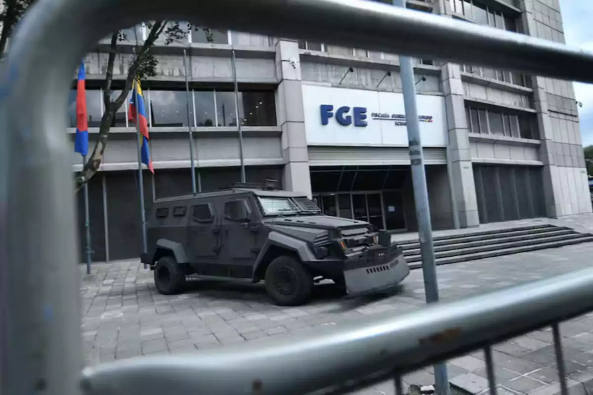 Armored vehicle parked in front of a government building with flags and a metal barrier in the foreground