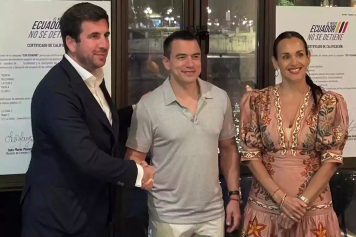 Tres personas posan para una foto en un evento formal, dos de ellas se dan la mano y al fondo hay carteles con el texto Ecuador no se detiene