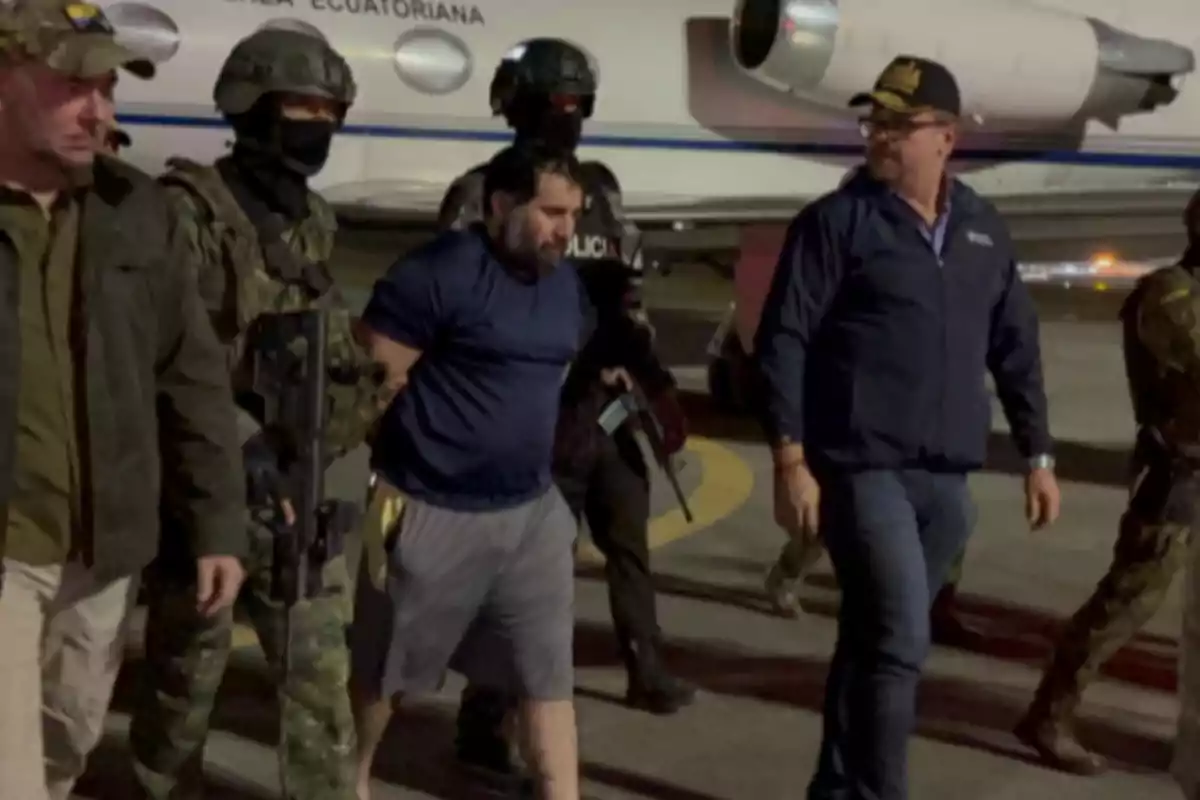 Un grupo de agentes escolta a un hombre esposado en la pista de un aeropuerto frente a un avión