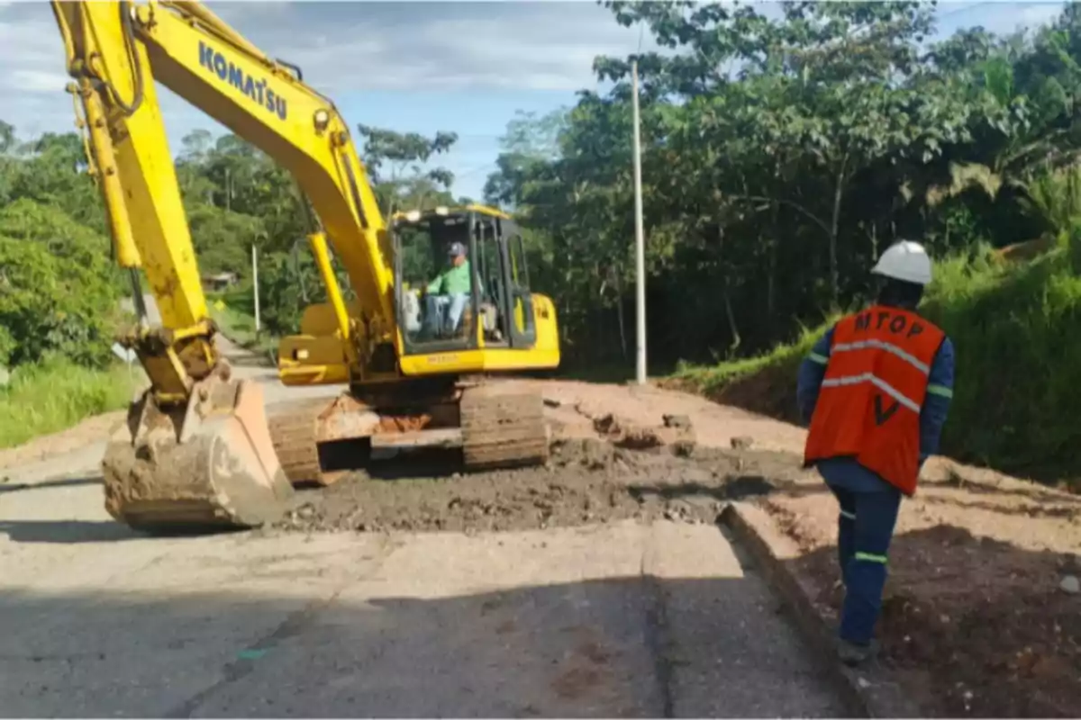 Yellow excavator working on road repairs while a worker wearing an orange vest and helmet supervises the work.