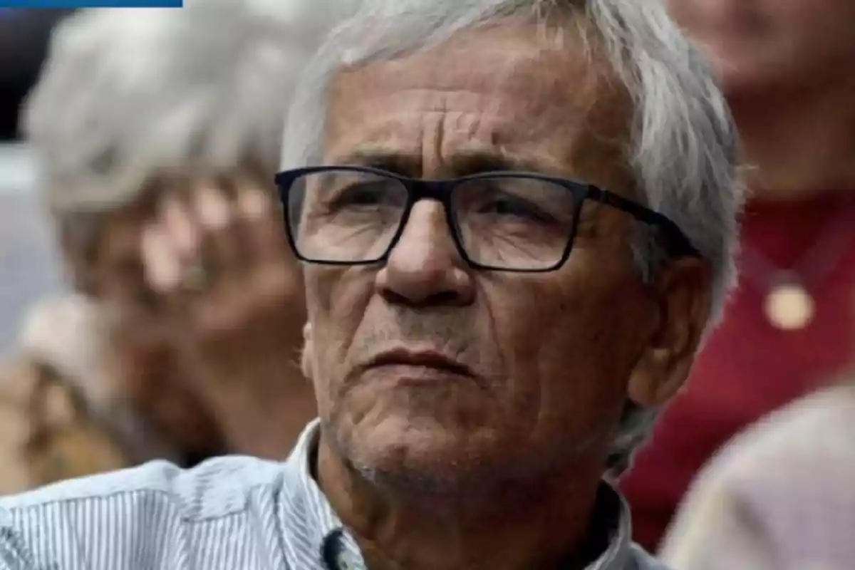 Hombre mayor de cabello canoso y gafas oscuras con expresión seria en un entorno interior Hombre mayor de cabello canoso y gafas oscuras con expresión seria en un entorno interior