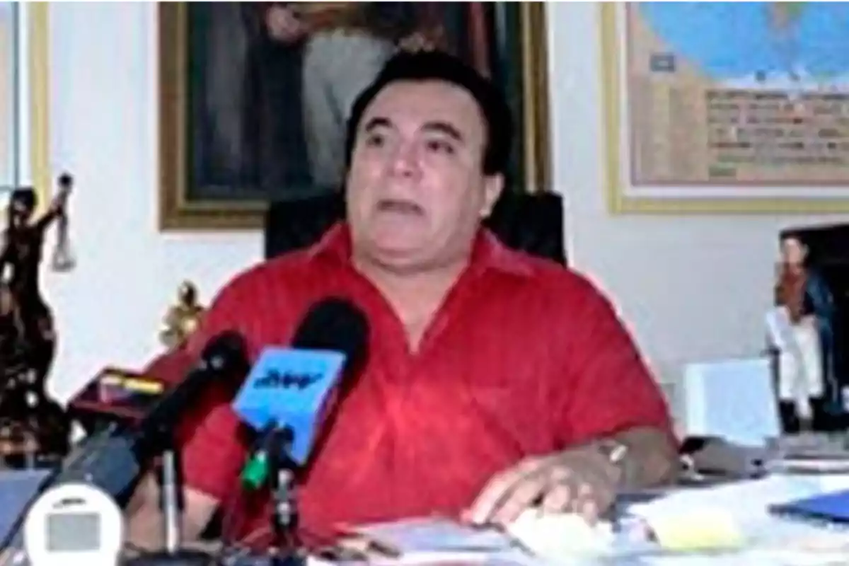 Man in a red jersey sitting at a desk with microphones in front of him and decorations in the background Man in a red jersey sitting at a desk with microphones in front of him and decorations in the background