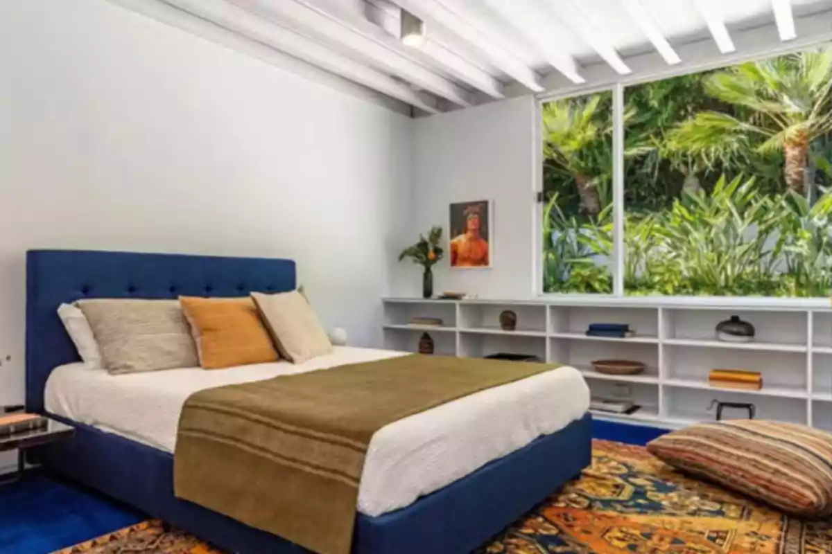 Modern bedroom with a blue headboard bed, neutral-toned cushions, brown blanket, colorful rug, white shelf with decorative objects, and large window overlooking green plants. Modern bedroom with a blue headboard bed, neutral-toned cushions, brown blanket, colorful rug, white shelf with decorative objects, and large window overlooking green plants.
