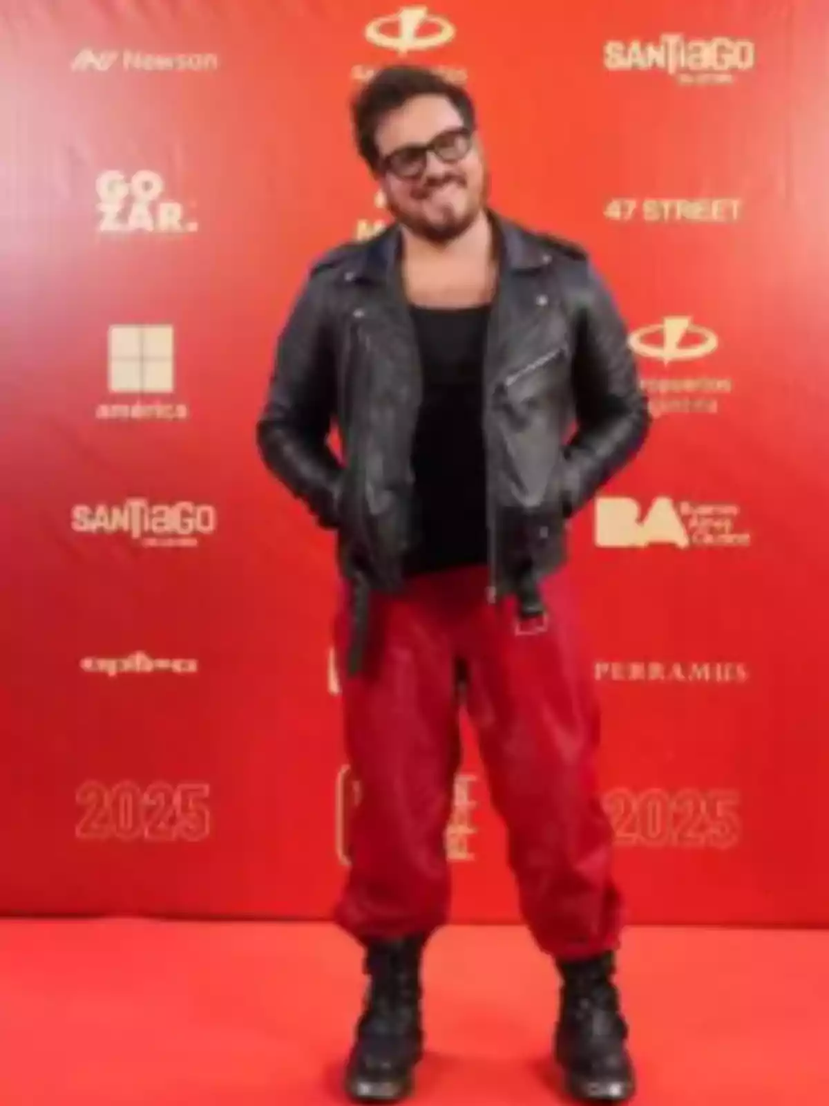 Persona posando sobre una alfombra roja con chaqueta de cuero negra, camiseta oscura, pantalones rojos y botas negras, frente a un fondo con logotipos y texto en color rojo Persona posando sobre una alfombra roja con chaqueta de cuero negra, camiseta oscura, pantalones rojos y botas negras, frente a un fondo con logotipos y texto en color rojo