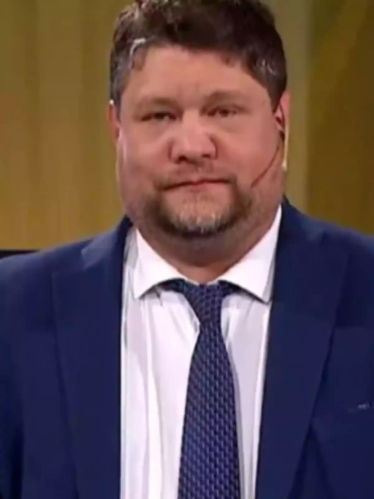 Hombre con barba y cabello corto usando traje azul, camisa blanca y corbata azul frente a un fondo amarillo Hombre con barba y cabello corto usando traje azul, camisa blanca y corbata azul frente a un fondo amarillo