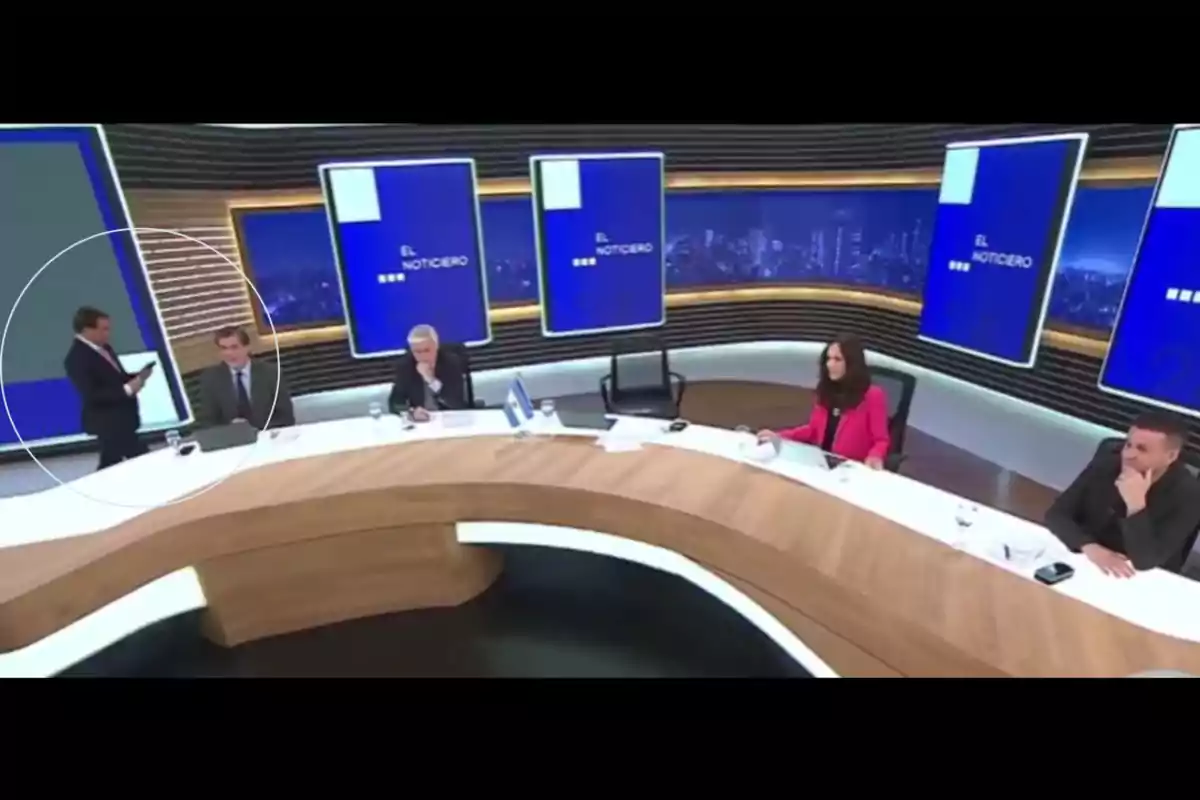 Cinco personas en un set de televisión con pantallas azules al fondo y una mesa curva de madera Cinco personas en un set de televisión con pantallas azules al fondo y una mesa curva de madera