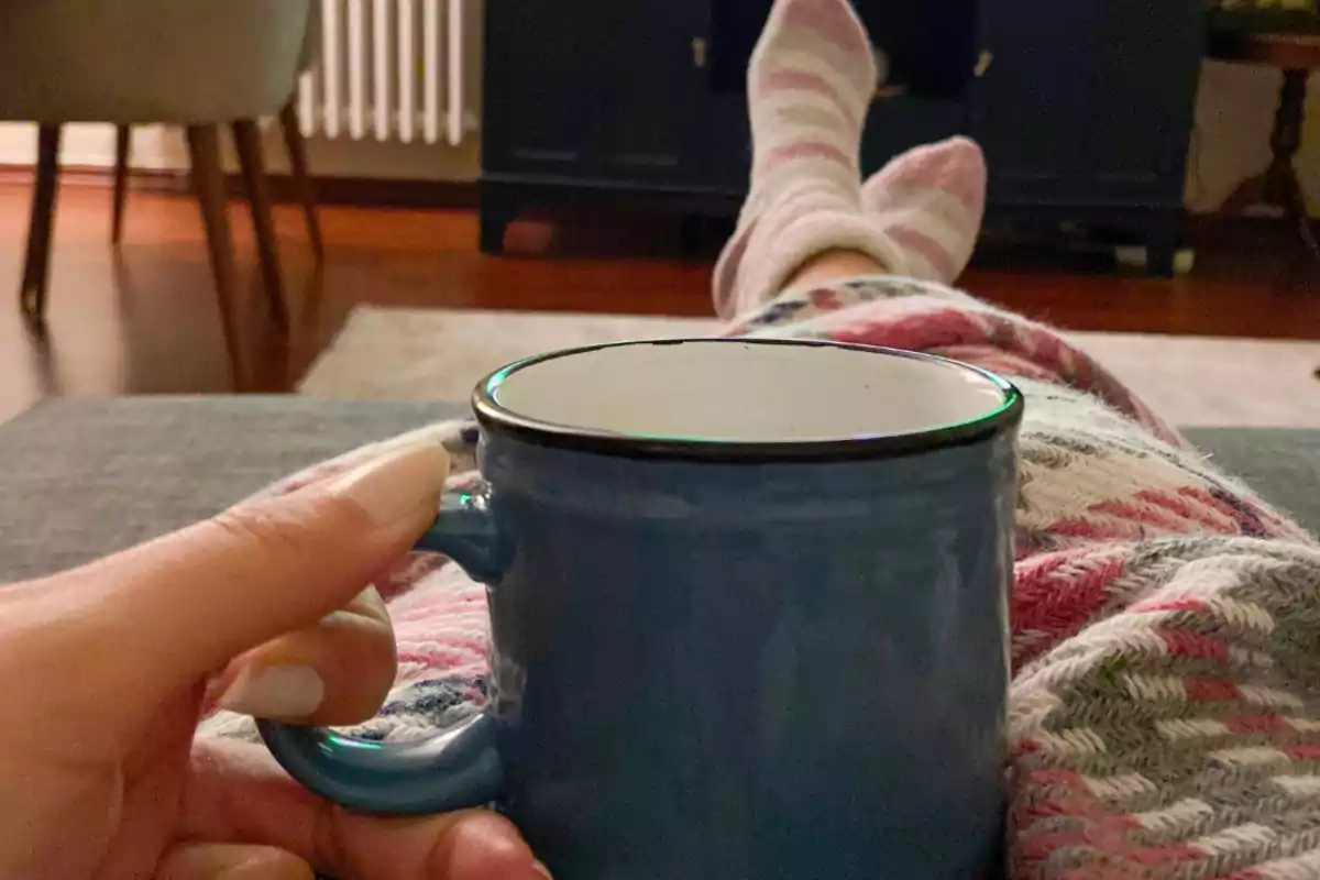 Persona recostada en un sofá con pijama y calcetas, sosteniendo una taza azul en una mano en un ambiente hogareño acogedor Persona recostada en un sofá con pijama y calcetas, sosteniendo una taza azul en una mano en un ambiente hogareño acogedor