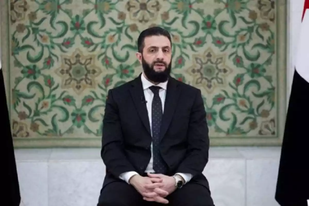 Hombre con barba y traje oscuro sentado frente a un fondo decorativo con patrones florales verdes y beige