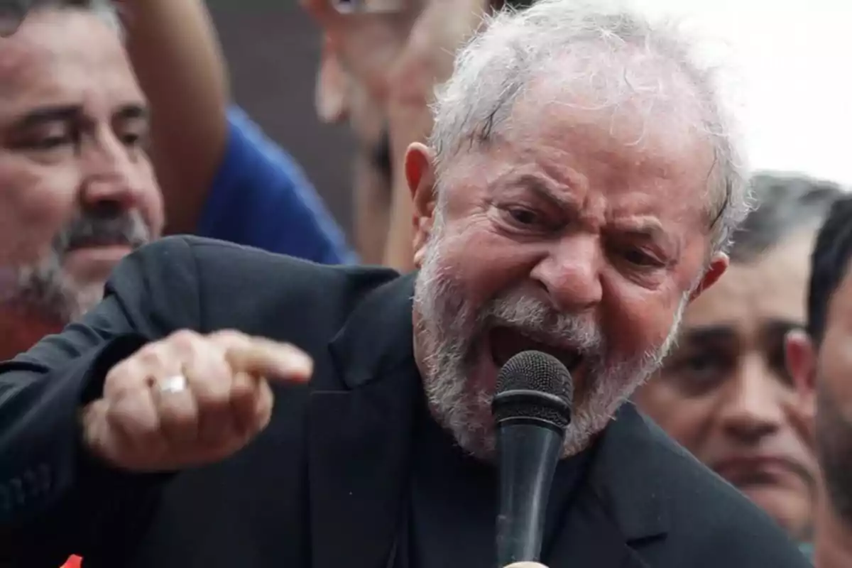 Hombre mayor con barba hablando apasionadamente por un micrófono y señalando con el dedo mientras varias personas lo rodean