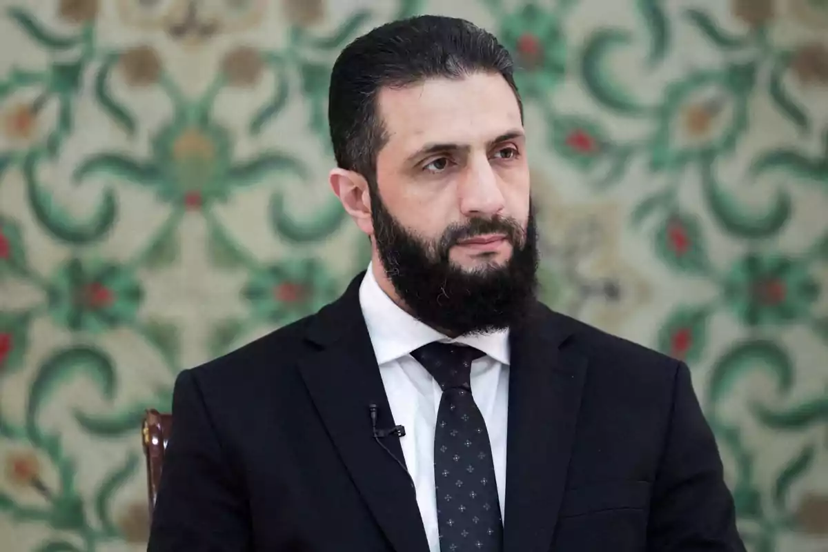 Hombre de barba oscura y cabello corto vestido con traje negro y corbata, sentado frente a un fondo decorativo con patrones verdes y rojos Hombre de barba oscura y cabello corto vestido con traje negro y corbata, sentado frente a un fondo decorativo con patrones verdes y rojos