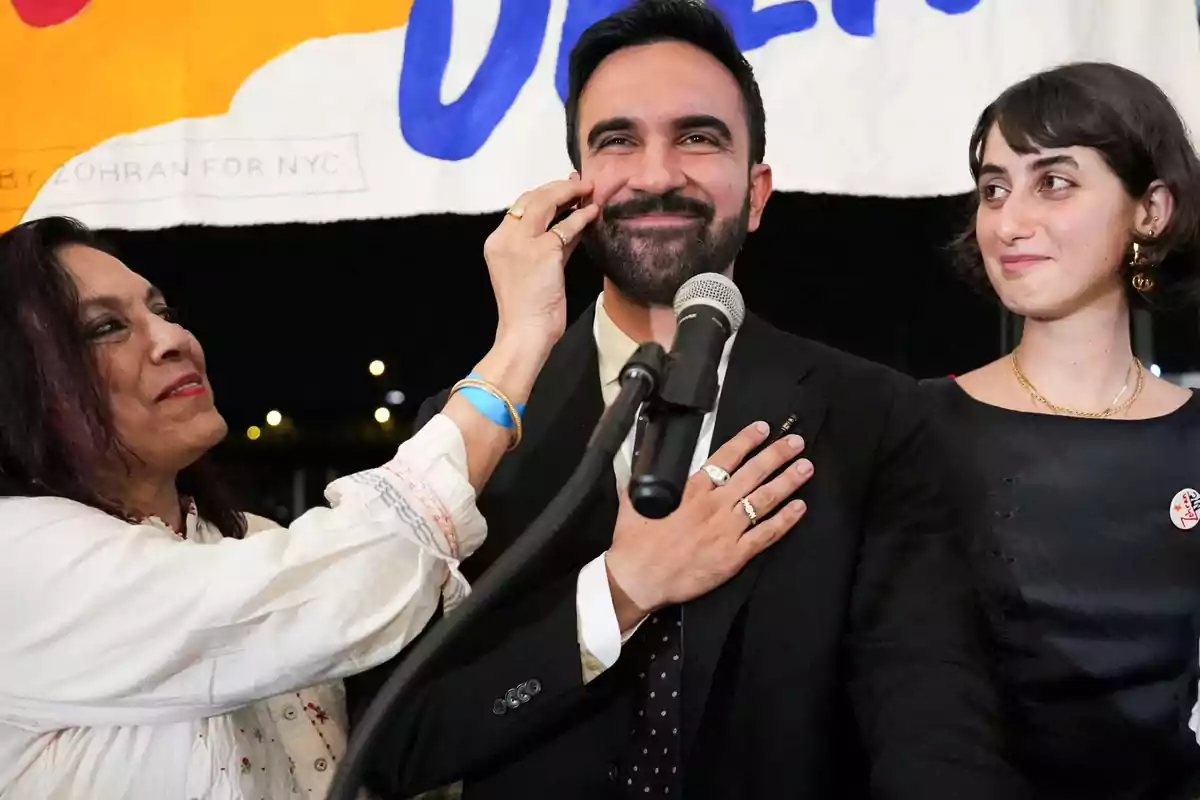 Una mujer toca la mejilla de un hombre sonriente que está frente a un micrófono, mientras otra mujer lo observa con una expresión alegre