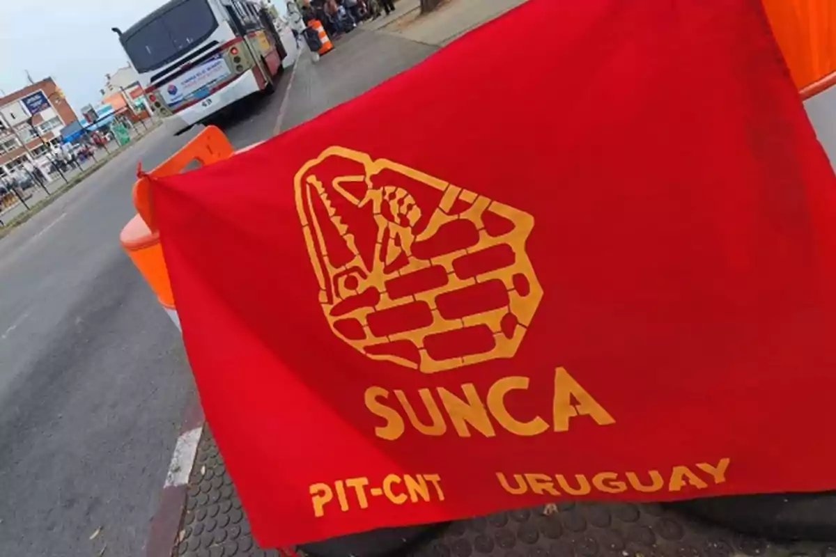 Bandera roja del sindicato SUNCA de Uruguay con un logo amarillo de un puño sosteniendo una herramienta y letras amarillas, en una calle con un autobús y personas al fondo Bandera roja del sindicato SUNCA de Uruguay con un logo amarillo de un puño sosteniendo una herramienta y letras amarillas, en una calle con un autobús y personas al fondo