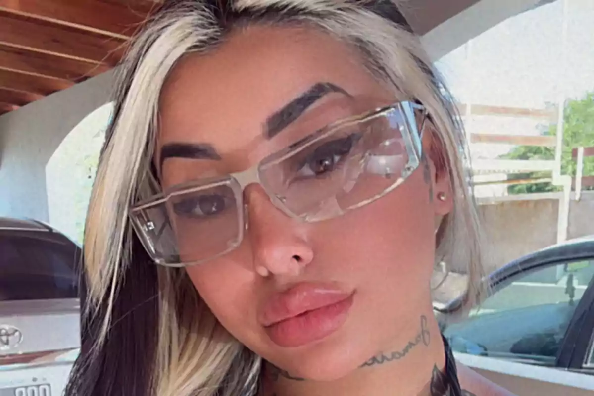Woman with large glasses and blonde and black hair posing outdoors next to parked cars