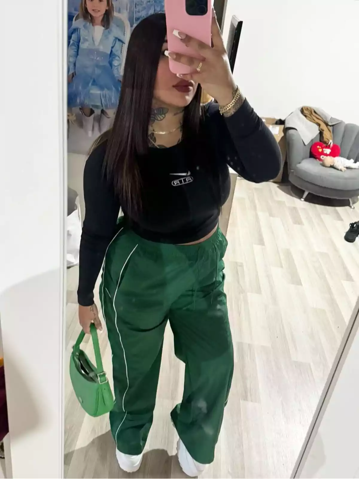 Mujer tomándose una selfie frente a un espejo usando pantalones verdes, top negro y bolso verde, con el fondo de una habitación moderna y un cuadro infantil en la pared.