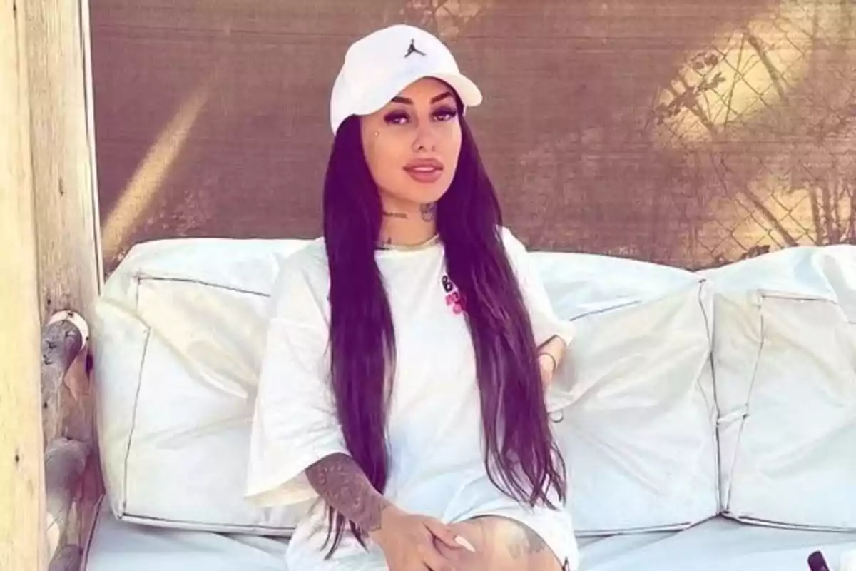 Mujer con cabello largo y oscuro sentada en un sofá blanco al aire libre, usando gorra blanca y camiseta blanca