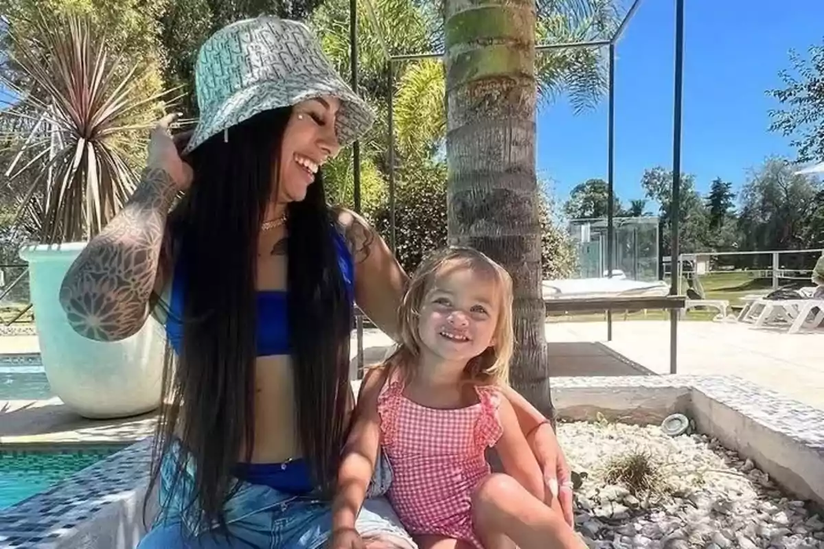 Mujer con sombrero y niña pequeña sonríen sentadas junto a una piscina en un día soleado rodeadas de palmeras y vegetación Mujer con sombrero y niña pequeña sonríen sentadas junto a una piscina en un día soleado rodeadas de palmeras y vegetación