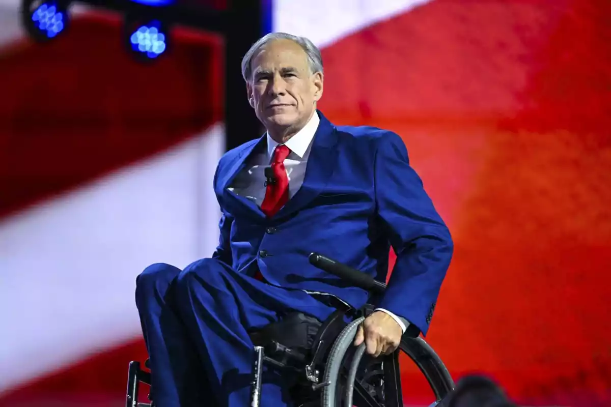Hombre de cabello canoso con traje azul y corbata roja sentado en una silla de ruedas frente a un fondo con colores rojo y blanco