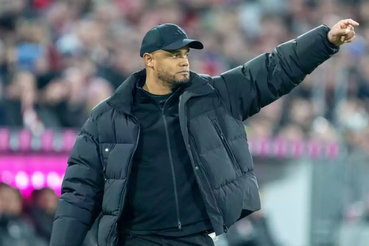 Hombre con chaqueta negra y gorra señalando hacia la derecha en un estadio lleno de gente Hombre con chaqueta negra y gorra señalando hacia la derecha en un estadio lleno de gente