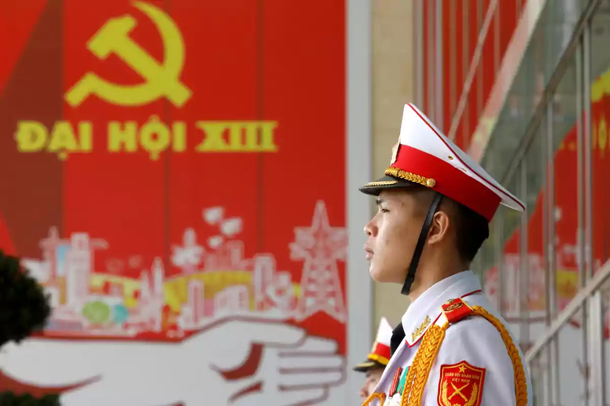 Soldado vietnamita con uniforme blanco y gorra roja de gala de pie frente a un cartel rojo con el símbolo comunista y letras amarillas Soldado vietnamita con uniforme blanco y gorra roja de gala de pie frente a un cartel rojo con el símbolo comunista y letras amarillas
