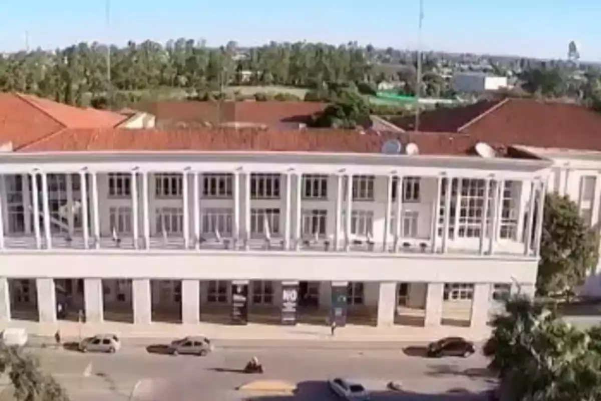 Edificio blanco de dos pisos con columnas y techo de tejas rojas ubicado junto a una calle por donde circulan varios autos Edificio blanco de dos pisos con columnas y techo de tejas rojas ubicado junto a una calle por donde circulan varios autos