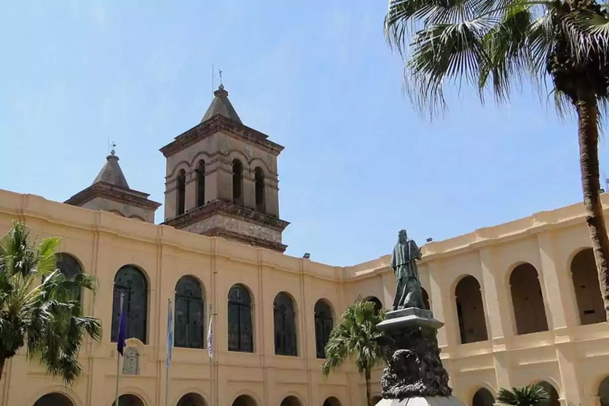 Edificio histórico de color claro con arcos y torres, rodeado de palmeras y una estatua en el centro del patio bajo un cielo despejado Edificio histórico de color claro con arcos y torres, rodeado de palmeras y una estatua en el centro del patio bajo un cielo despejado