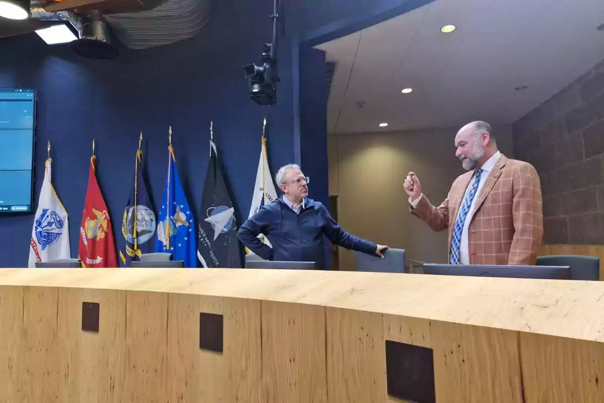 Dos hombres conversan en una sala de conferencias con varias banderas militares al fondo y una mesa de madera en primer plano Dos hombres conversan en una sala de conferencias con varias banderas militares al fondo y una mesa de madera en primer plano