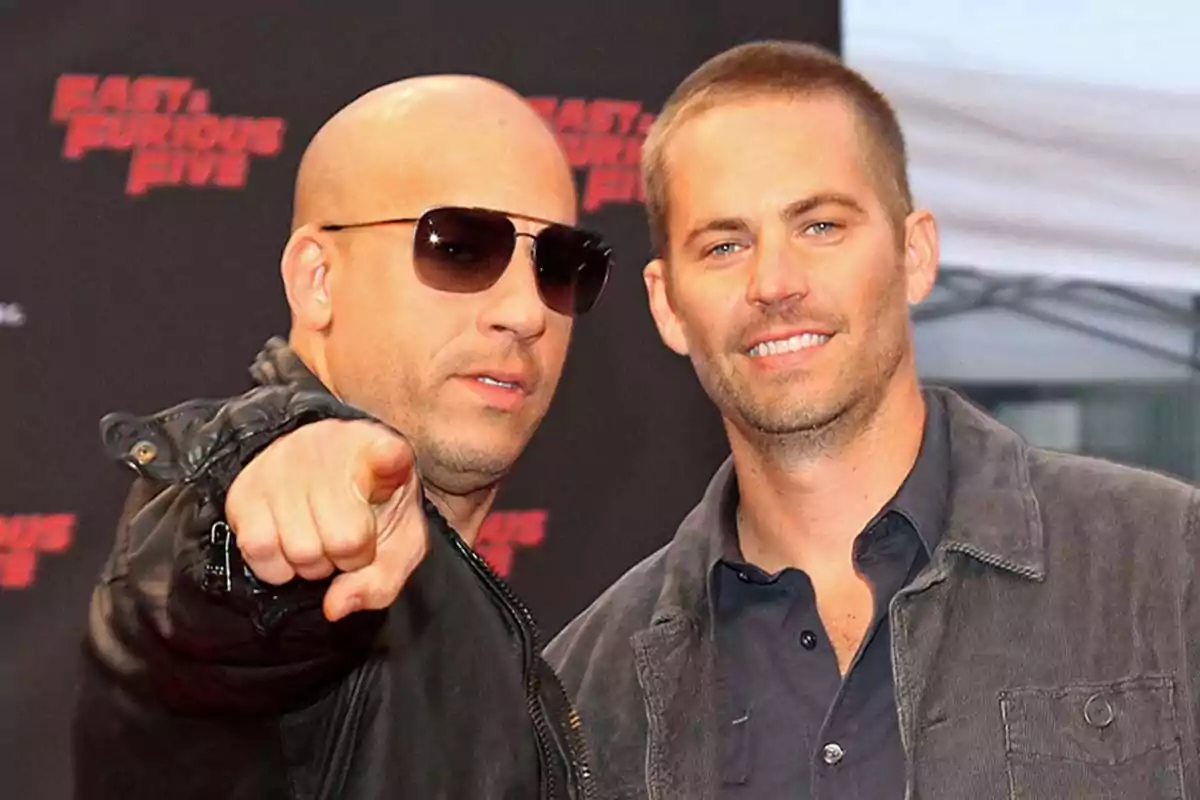 Dos hombres posan juntos en un evento, uno de ellos lleva gafas de sol y señala hacia la cámara mientras el otro sonríe