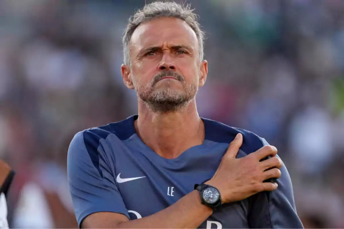 Short-haired, gray-haired man with a beard, wearing a blue sports jersey and a black watch, with a serious expression and his right hand on his left shoulder in a blurred outdoor setting. Short-haired, gray-haired man with a beard, wearing a blue sports jersey and a black watch, with a serious expression and his right hand on his left shoulder in a blurred outdoor setting.