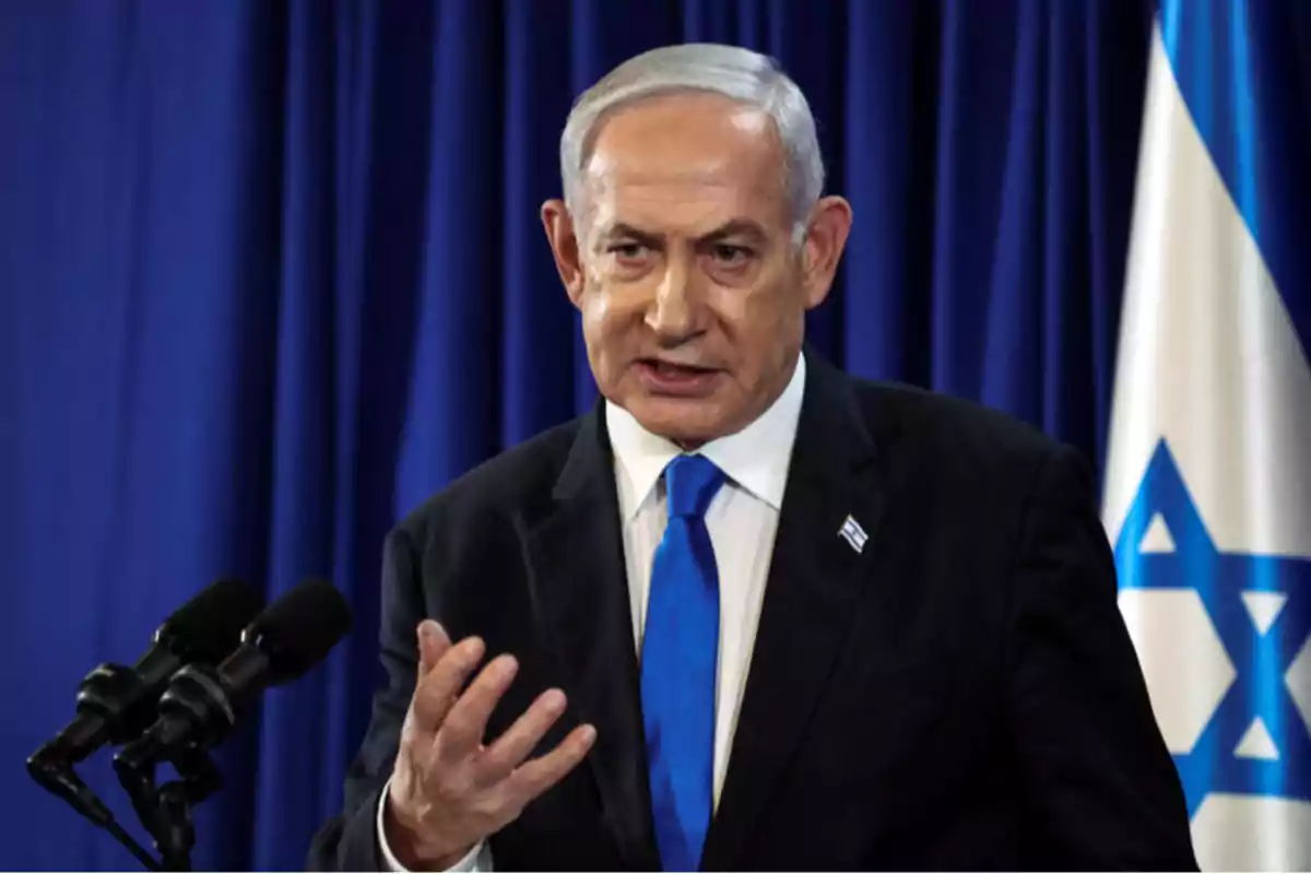 Gray-haired man in a dark suit and blue tie speaking in front of microphones with an Israeli flag in the background Gray-haired man in a dark suit and blue tie speaking in front of microphones with an Israeli flag in the background