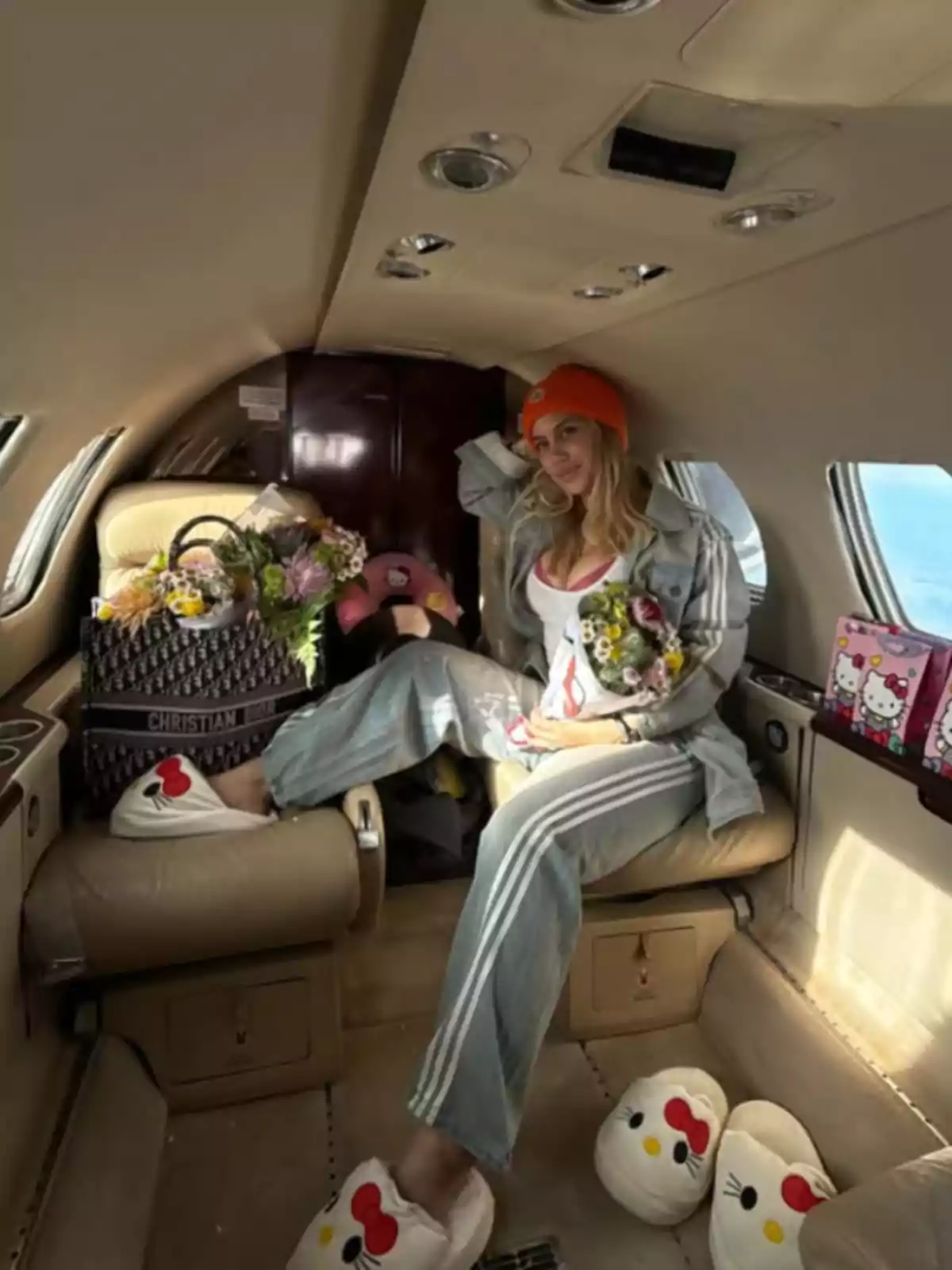 Mujer sentada en un jet privado usando pantuflas de Hello Kitty y gorro naranja, rodeada de flores y una bolsa de diseñador Mujer sentada en un jet privado usando pantuflas de Hello Kitty y gorro naranja, rodeada de flores y una bolsa de diseñador