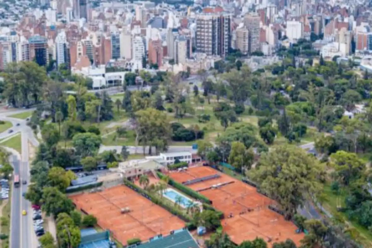 Vista aérea de canchas de tenis de polvo de ladrillo rodeadas de árboles y áreas verdes con una ciudad de fondo Vista aérea de canchas de tenis de polvo de ladrillo rodeadas de árboles y áreas verdes con una ciudad de fondo