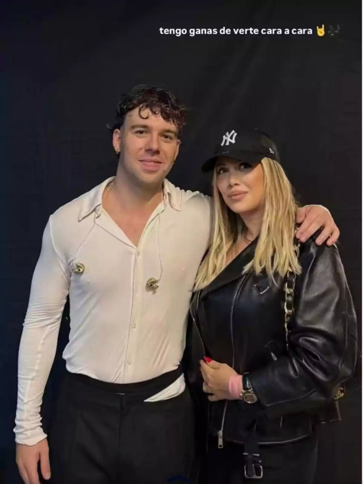 Dos personas posan juntas frente a un fondo oscuro, una de ellas lleva una camisa blanca y la otra una chaqueta de cuero negra y gorra negra Dos personas posan juntas frente a un fondo oscuro, una de ellas lleva una camisa blanca y la otra una chaqueta de cuero negra y gorra negra