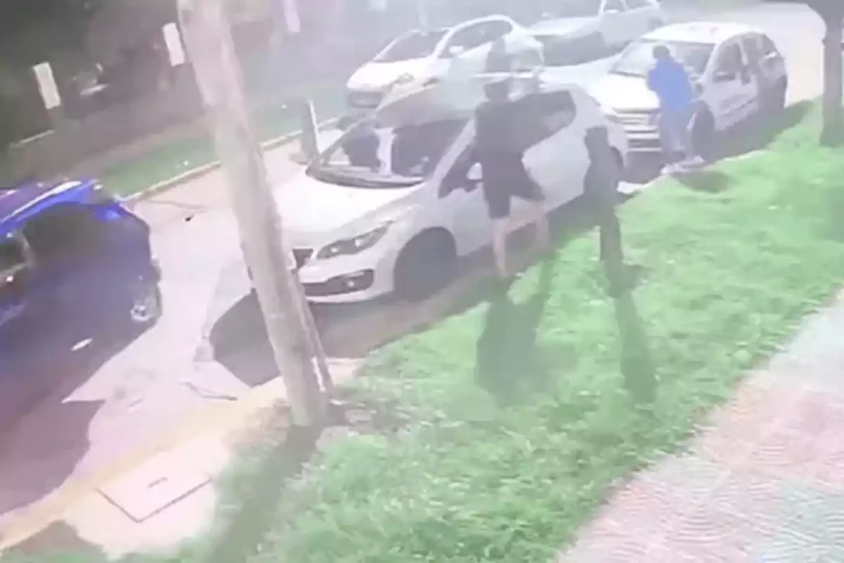 Three people next to a white car parked on the street at night, one of them appears to be forcing the vehicle’s door while another watches and a third individual is near another white car further ahead. Three people next to a white car parked on the street at night, one of them appears to be forcing the vehicle’s door while another watches and a third individual is near another white car further ahead.