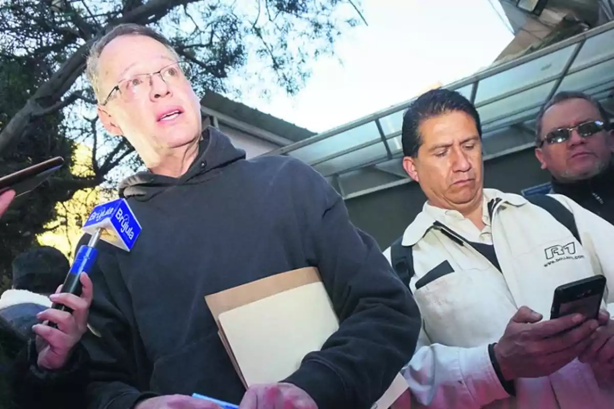 Un hombre con gafas y sudadera oscura sostiene una carpeta mientras es entrevistado por reporteros, uno de ellos sostiene un micrófono azul y otro revisa su teléfono móvil, al fondo hay árboles y una estructura metálica. Un hombre con gafas y sudadera oscura sostiene una carpeta mientras es entrevistado por reporteros, uno de ellos sostiene un micrófono azul y otro revisa su teléfono móvil, al fondo hay árboles y una estructura metálica.