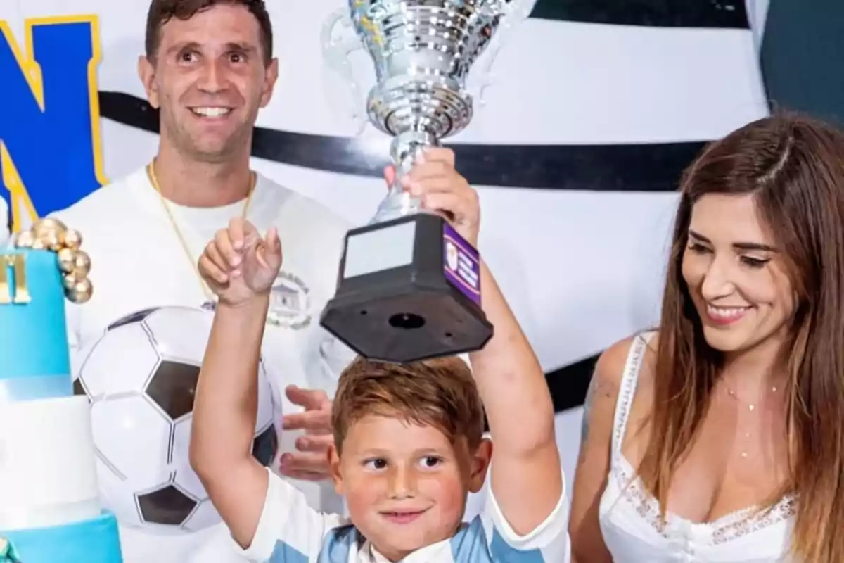 A smiling boy lifts a silver trophy while a man and a woman watch him happily at a soccer-themed celebration.