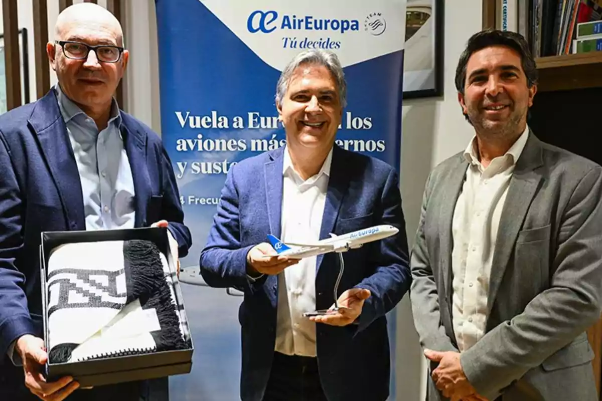 Tres hombres posan sonrientes en un evento corporativo de Air Europa uno sostiene una caja con una manta y otro una maqueta de avión frente a un cartel promocional de la aerolínea Tres hombres posan sonrientes en un evento corporativo de Air Europa uno sostiene una caja con una manta y otro una maqueta de avión frente a un cartel promocional de la aerolínea
