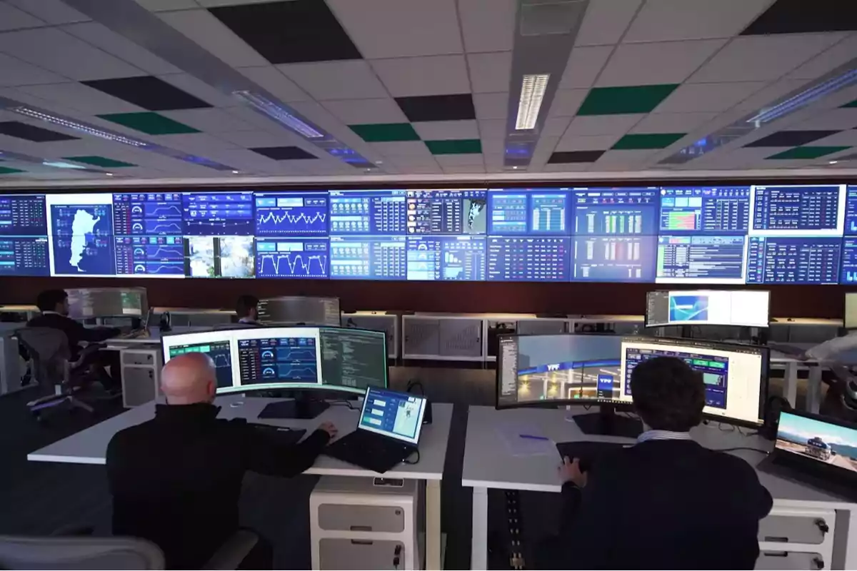 Personas trabajando en una sala de control con múltiples monitores y pantallas grandes mostrando gráficos y datos en tiempo real