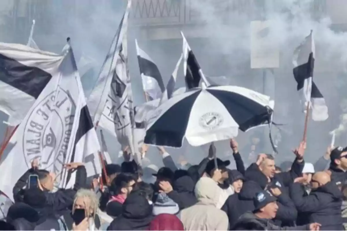 Aficionados reunidos en una manifestación deportiva ondeando banderas y sosteniendo un paraguas blanco y negro entre humo