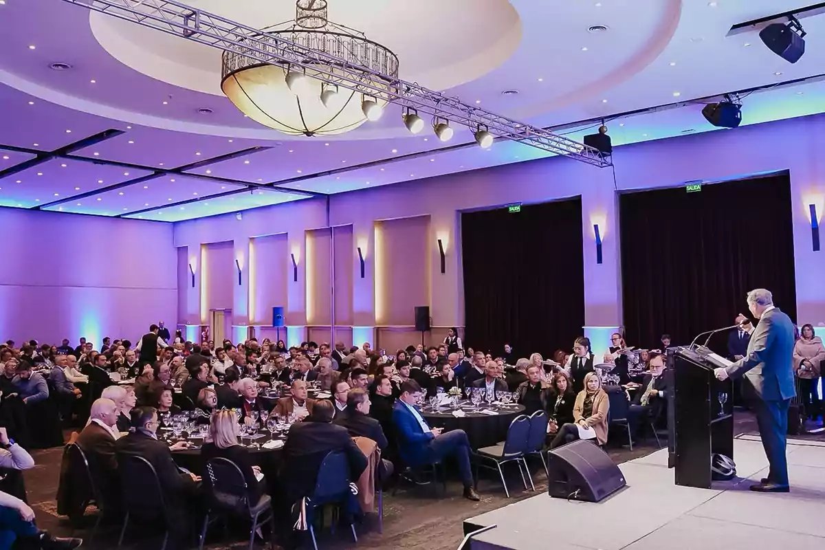 Un hombre de traje habla en un podio frente a una audiencia numerosa sentada en mesas redondas dentro de un salón elegante iluminado con luces azules