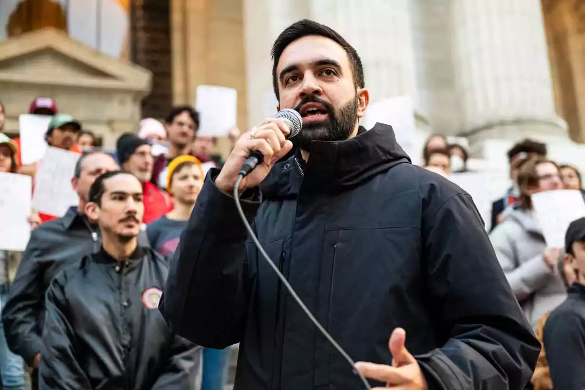 Hombre con barba y chaqueta negra hablando por micrófono frente a un grupo de personas manifestándose con carteles en la mano Hombre con barba y chaqueta negra hablando por micrófono frente a un grupo de personas manifestándose con carteles en la mano
