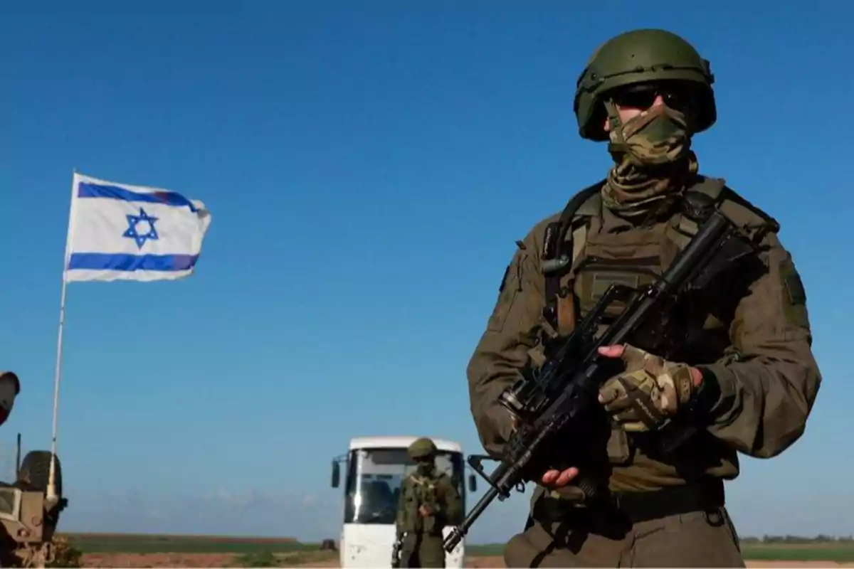 Soldado con uniforme militar y arma en primer plano, bandera de Israel ondeando al fondo junto a un vehículo blanco en un paisaje despejado