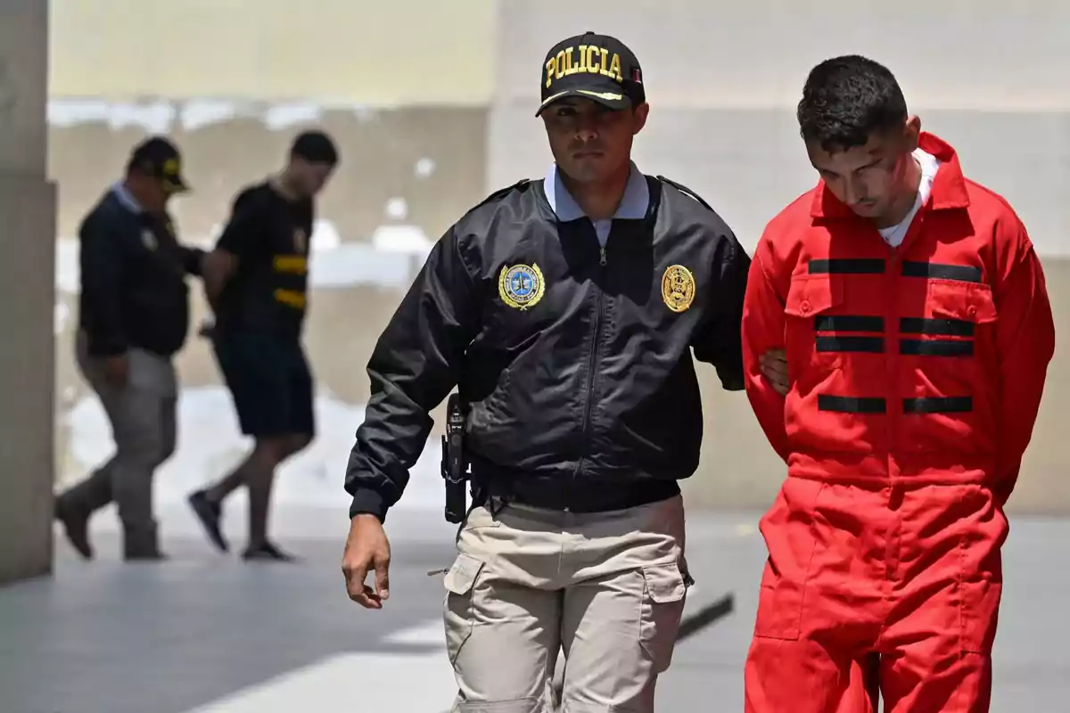 Un policía escolta a un hombre con uniforme rojo mientras al fondo se observa a otro agente con una persona detenida. Un policía escolta a un hombre con uniforme rojo mientras al fondo se observa a otro agente con una persona detenida.