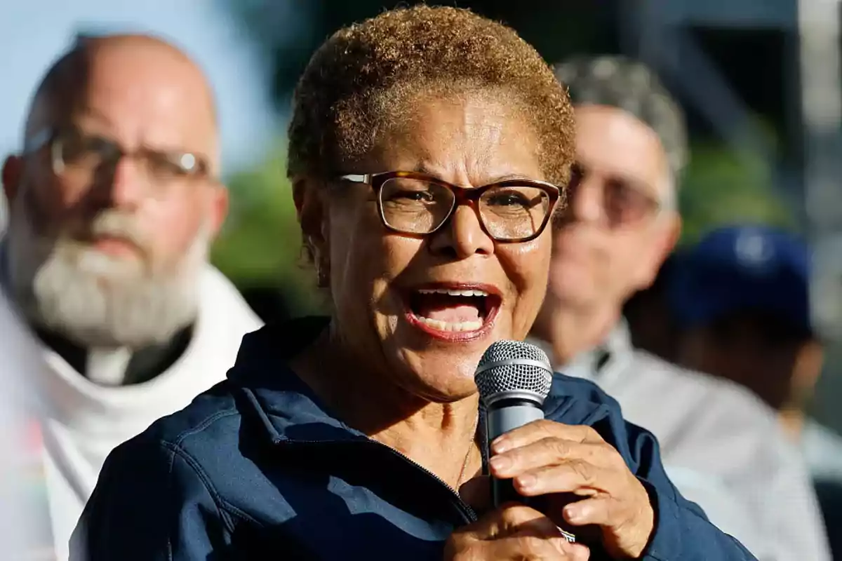 Short-haired woman with glasses speaking into a microphone during an outdoor event with blurred people in the background Short-haired woman with glasses speaking into a microphone during an outdoor event with blurred people in the background