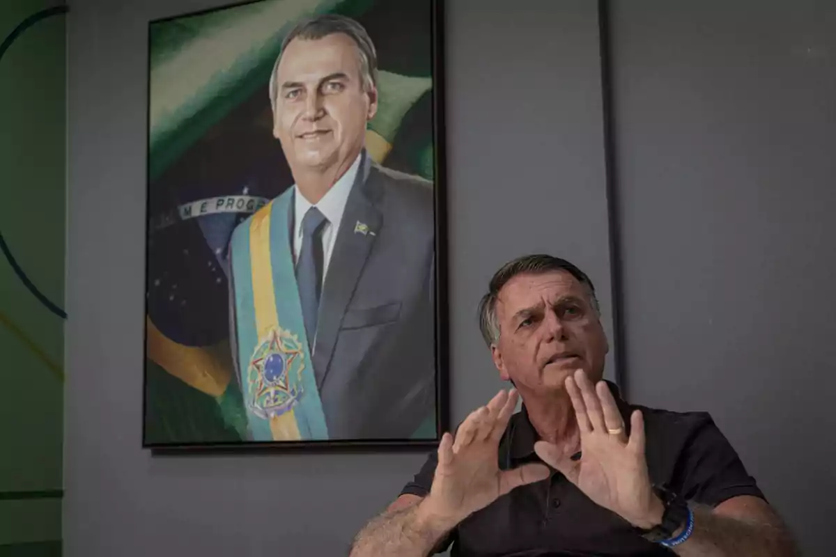 Un hombre sentado gesticulando con las manos frente a un retrato suyo en la pared, ambos con expresión seria.