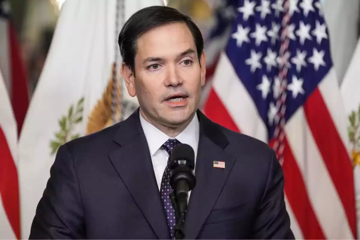 Hombre de cabello oscuro con traje y corbata hablando frente a un micrófono con banderas de Estados Unidos y México al fondo Hombre de cabello oscuro con traje y corbata hablando frente a un micrófono con banderas de Estados Unidos y México al fondo