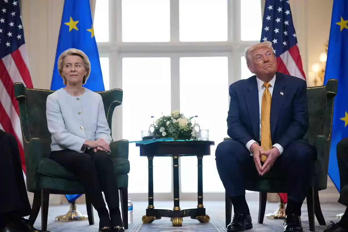 Two people sitting in green armchairs with European Union and United States flags in the background and a small table with flowers between them