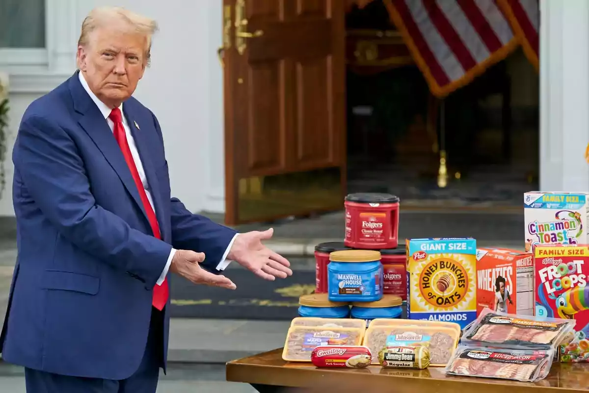 Un hombre de traje azul y corbata roja muestra una mesa llena de productos alimenticios como cereales, café y tocino frente a una puerta abierta y una bandera estadounidense. Un hombre de traje azul y corbata roja muestra una mesa llena de productos alimenticios como cereales, café y tocino frente a una puerta abierta y una bandera estadounidense.