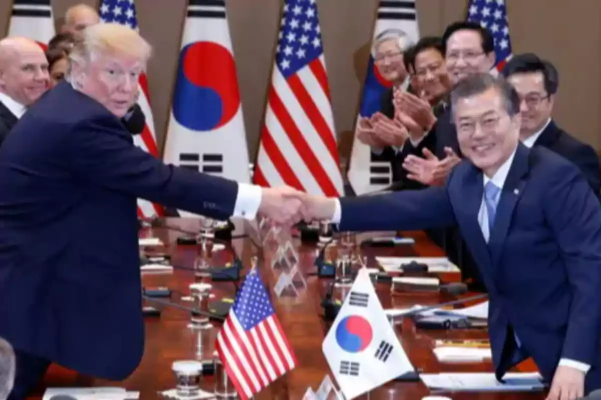 Dos hombres de traje se dan la mano en una mesa de reuniones rodeados de banderas de Estados Unidos y Corea del Sur mientras varias personas aplauden al fondo Dos hombres de traje se dan la mano en una mesa de reuniones rodeados de banderas de Estados Unidos y Corea del Sur mientras varias personas aplauden al fondo